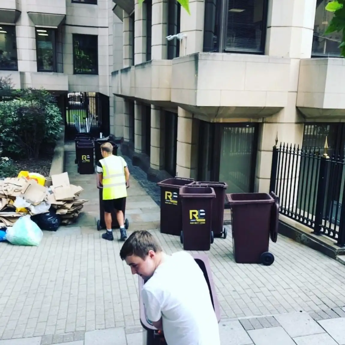 Two RLE crew members moving RLE-branded wheelie bins outside an office, with flattened cardboard and bags ready to load