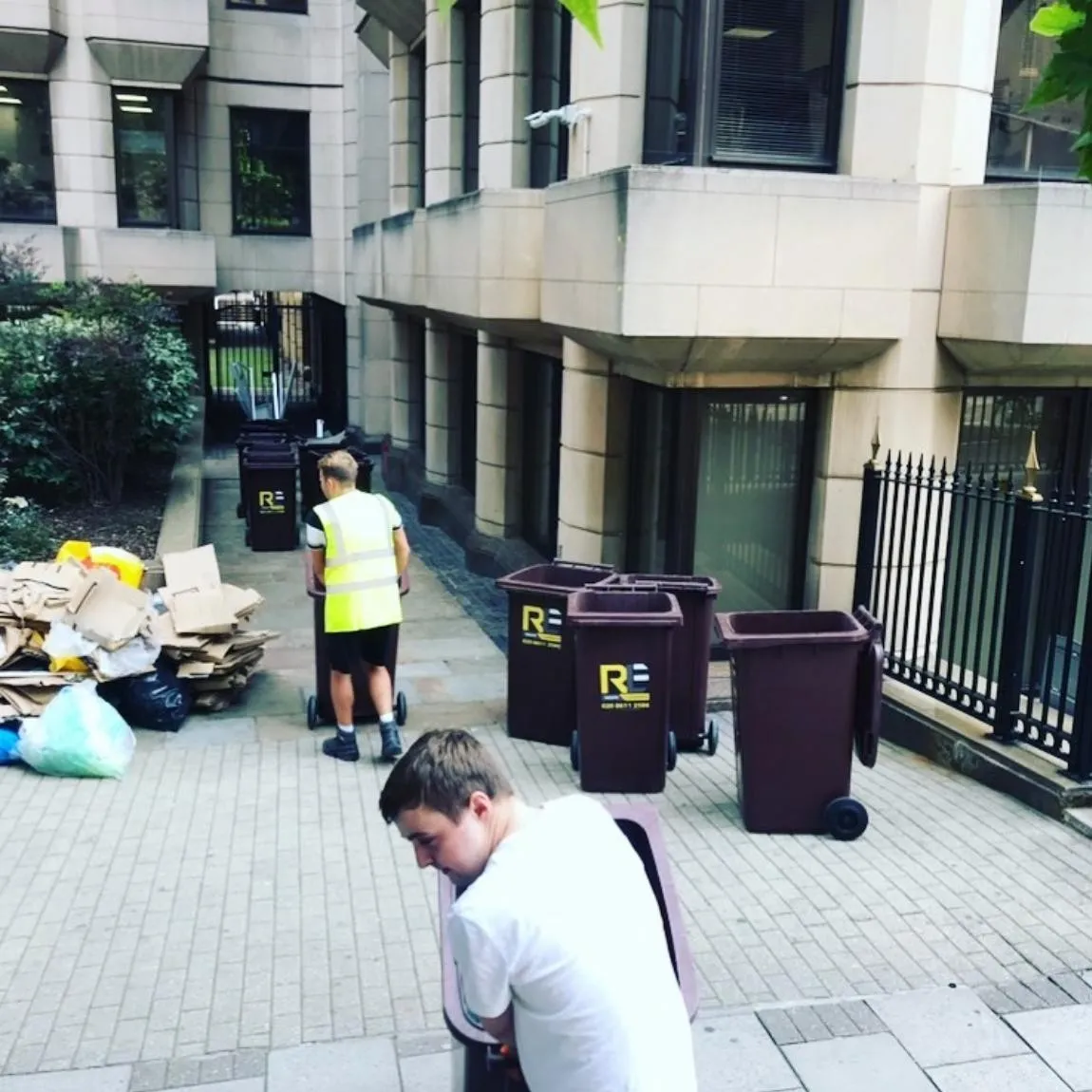 Two RLE crew members, one in hi-vis, moving RLE wheelie bins outside an office, with flattened cardboard and bags to load