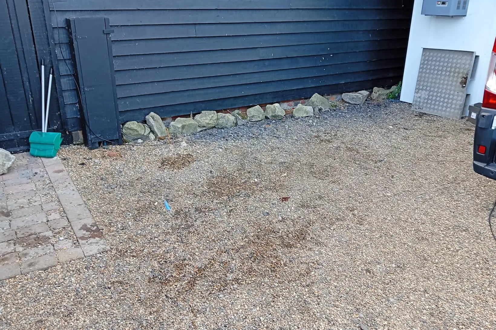 After: The same gravel yard cleared, with a board leaning on the timber-clad wall and a row of stones along its base