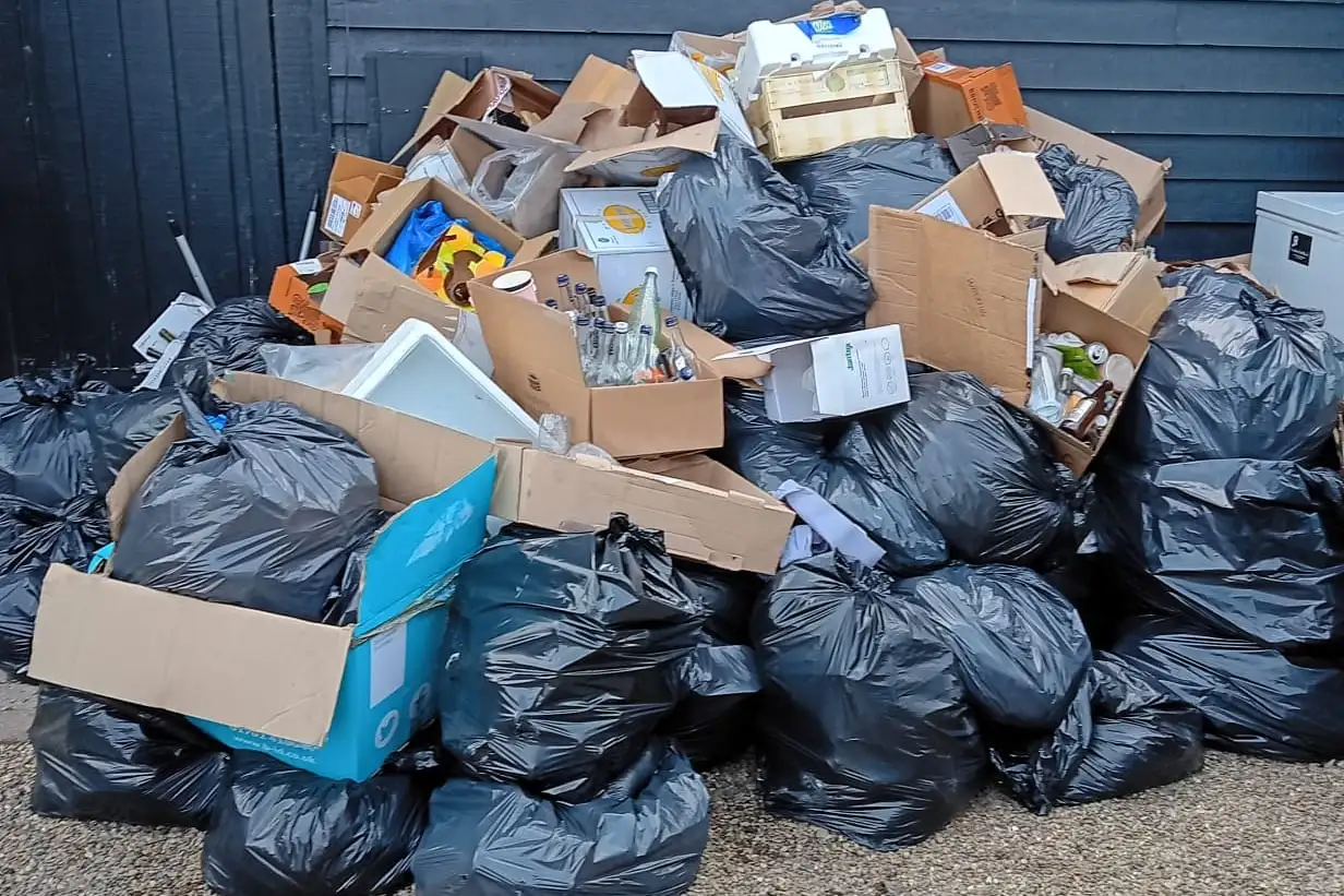 Black bin bags and cardboard boxes of rubbish piled high against a black timber-clad wall on a gravel yard