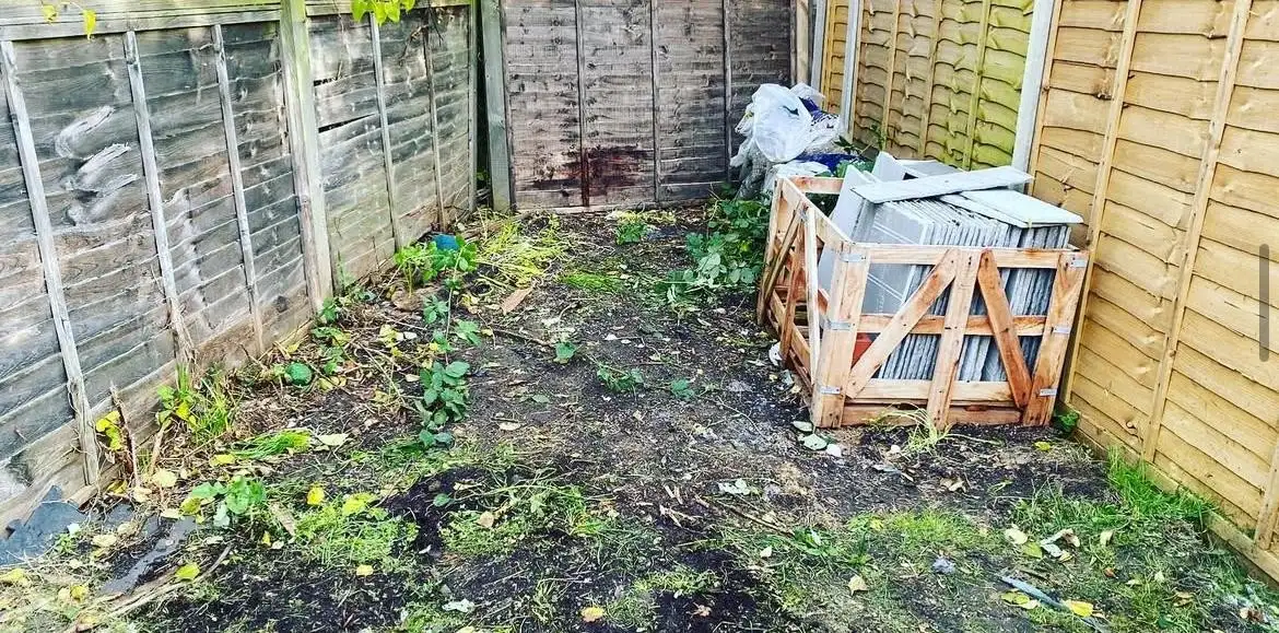 After: The same strip of garden cleared to bare earth between wooden fences, with only the crate of paving slabs left