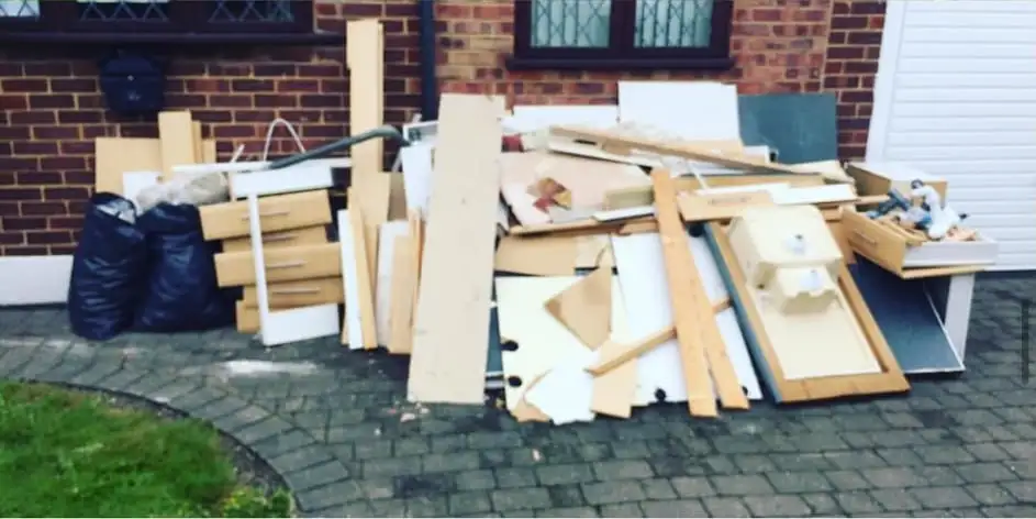 Old kitchen cabinets, worktops and boards stacked with black bags on a block-paved drive in front of a brick house